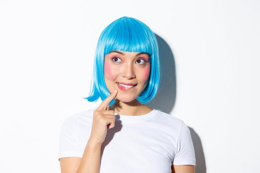 Image of thoughtful cute asian girl in blue wig, looking at upper left corner and thinking, standing over white background.