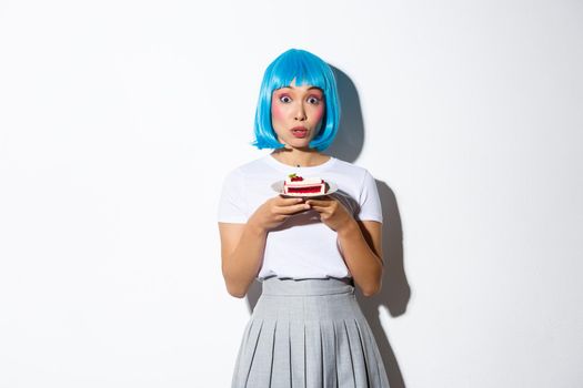 Portrait of surprised silly asian girl looking coquettish at camera, holding cake on plate, wearing blue anime wig, standing over white background.
