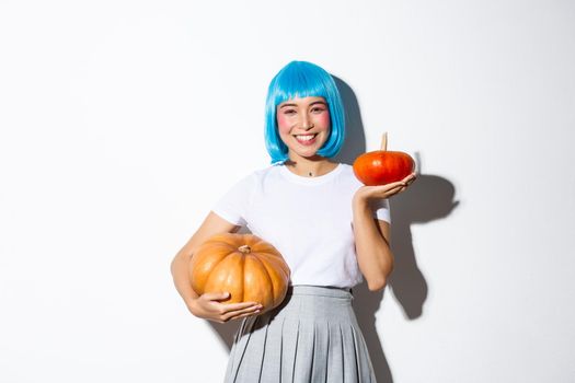 Image of adorable asian girl in blue wig, celebrating halloween, showing large and small pumpkins and smiling happy, standing over white background.