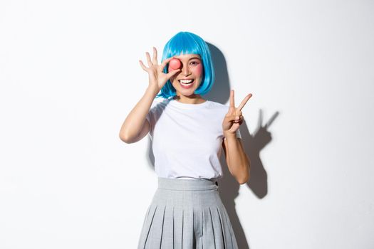 Beautiful japanese girl in blue wig and schoolgirl costume holding macaron, showing peace gesture and smiling, standing over white background.