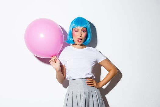 Image of flirty asian woman winking and showing tongue, wearing blue wig for halloween party, holding large pink balloon, standing over white background.