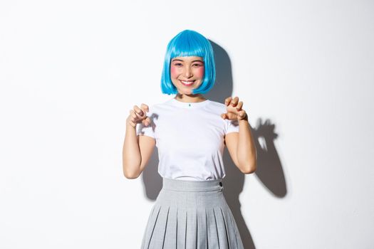 Funny and cute asian girl in blue wig smiling and making hand paws gesture, celebrating halloween, standing over white background.