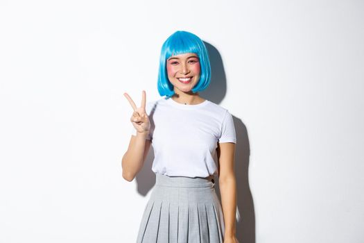 Image of beautiful anime girl in blue wig showing peace gesture. Woman wearing outfit for halloween, smiling happy and standing over white background.