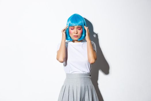 Portrait of concerned young asian woman close eyes and touching head, suffering headache or migraine, dressed-up for halloween party, standing over white background.