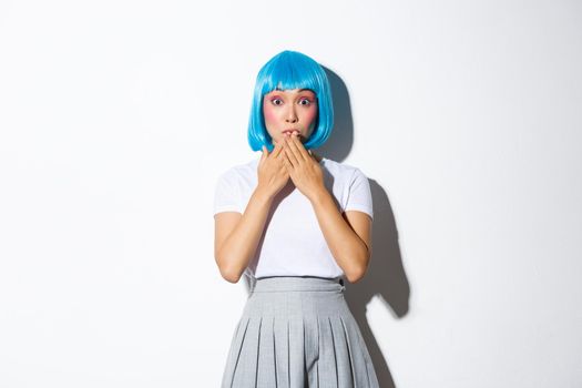 Portrait of surprised beautiful asian girl in glamour blue wig, looking amazed, holding hands near lips thrilled, standing over white background.