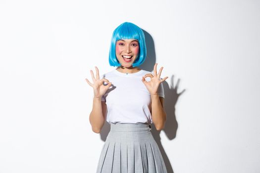 Portrait of satisfied excited girl in blue wig and halloween outfit showing okay gesture, approve something or recommend, standing over white background.