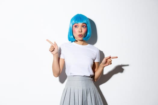 Image of amazed asian girl in blue wig turn attention to advertisements, pointing fingers sideways and looking amazed, standing over white background.