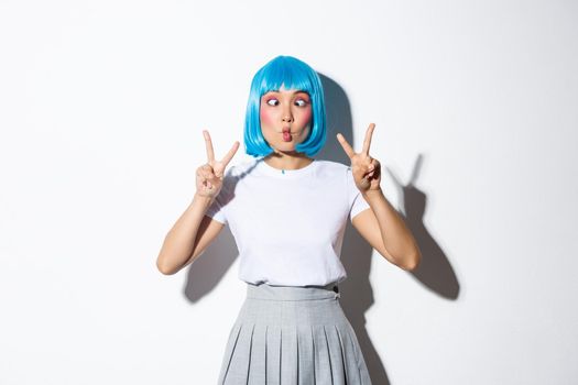 Portrait of silly and cute asian girl in blue wig, squinting and making funny faces, showing peace gestures, standing over white background.