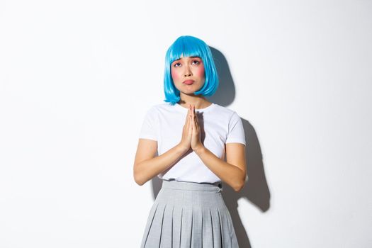 Portrait of hopeful sad girl looking up and praying or begging for God help, standing in blue wig over white background.
