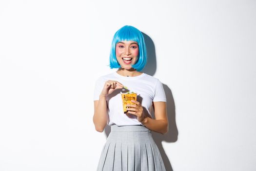 Image of cute asian girl celebrating halloween, eating sweets from trick or treat bag, wearing blue short wig and anime schoolgirl costume, standing over white background.