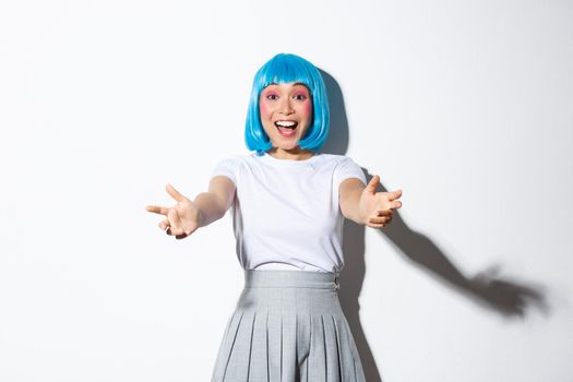 Portrait of happy asian girl in blue short wig and schoolgirl outfit stretching hands forward to hold or catch something, reaching for hug and smiling, standing over white background.