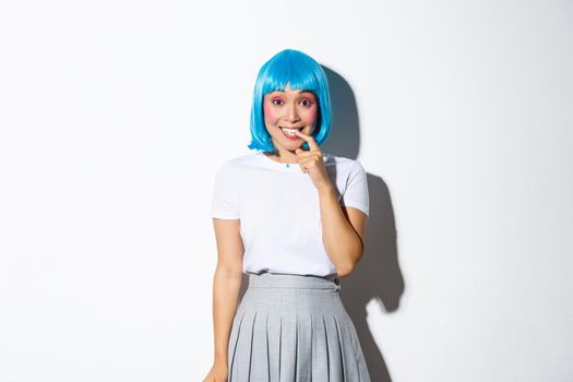 Portrait of silly asian girl in blue wig, smiling and looking curious, standing over white background tempted.