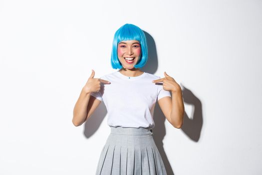 Portrait of beautiful korean girl in blue wig pointing at herself with cheerful smile, standing over white background in halloween costume.