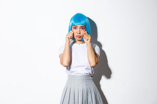 Image of sad asian girl in blue wig and schoolgirl uniform, dressed-up for halloween party, crying and wiping tears, feeling upset, standing over white background.