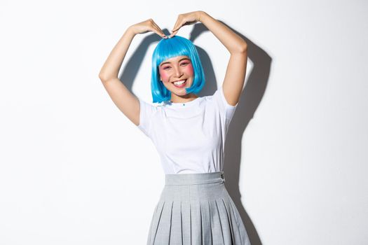 Lovely asian girl celebrating halloween, wearing blue wig and school uniform, showing heart gesture and smiling, standing over white background.