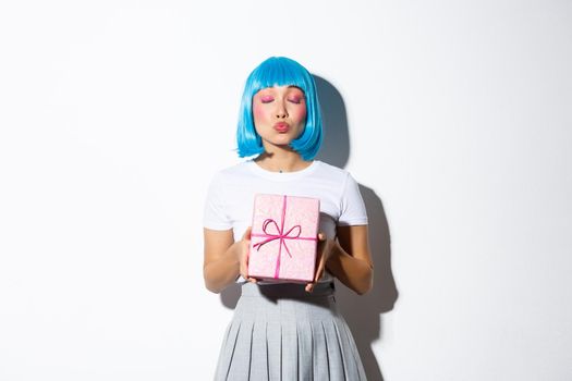 Beautiful asian girl in blue party wig kissing someone and giving a gift wrapped in pink paper, standing over white background.