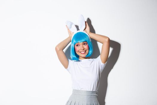 Portrait of cute smiling asian girl in blue short wig and rabbit ears, looking silly at camera, wearing halloween party costume, standing over white background.