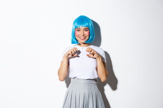 Portrait of beautiful asian girl in blue wig, enjoying halloween, smiling and having fun at party, standing over white background.