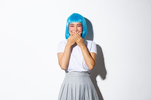 Portrait of cute and silly asian girl having fun, giggle with hands pressed to mouth, standing over white background, enjoying halloween party, wearing blue wig and school uniform.