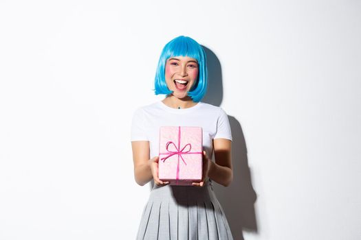Image of cheerful cute asian girl in blue party wig, giving you a gift wrapped in pink paper, congratulating with something, standing over white background.