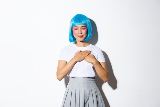 Portrait of lovely asian girl in blue stylish wig, close eyes and smiling while holding hands on heart and recall something, daydreaming over white background.