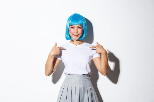 Beautiful asian girl in blue wig pointing fingers at herself and smiling, standing confident over white background, wearing halloween costume.