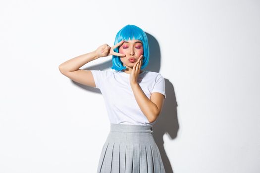 Portrait of lovely asian girl in blue wig close eyes and pouting for kiss, showing kawaii peace gesture, standing over white background.