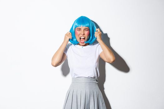 Portrait of outraged asian woman in blue wig looking mad, yelling at someone, standing over white background.