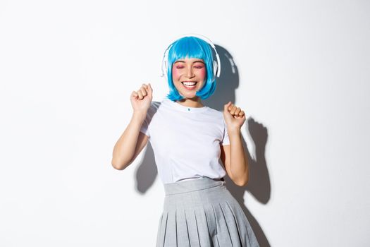 Portrait of happy asian girl in blue wig dancing, listening music in headphones over white background.