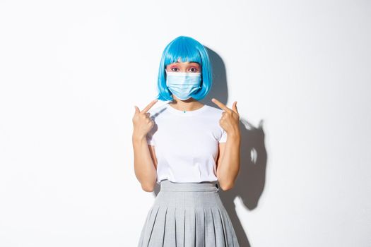 Concept of halloween celebration and covid-19 pandemic. Image of asian girl in blue wig pointing at medical mask, standing over white background.