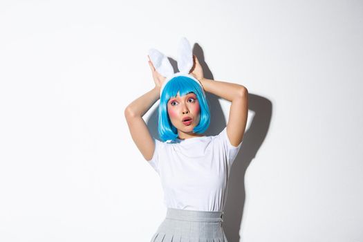 Image of pretty asian girl looking surprised left, wearing blue wig and rabbit ears for halloween party, standing over white background.