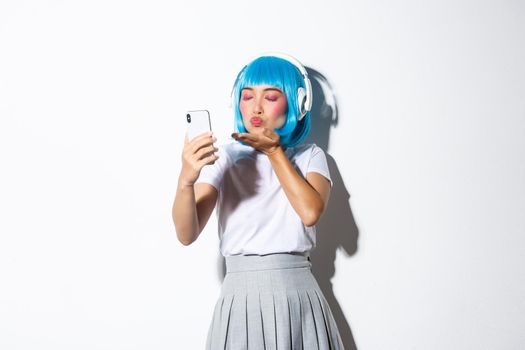 Portrait of cute asian girl dressed up as anime character, posing for selfie in headphones and blue short wig, taking picture on smartphone, standing over white background.