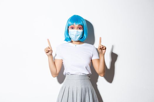 Concept of halloween celebration and covid-19 pandemic. Happy asian girl in blue wig and medical mask, showing advertisement, pointing fingers up.