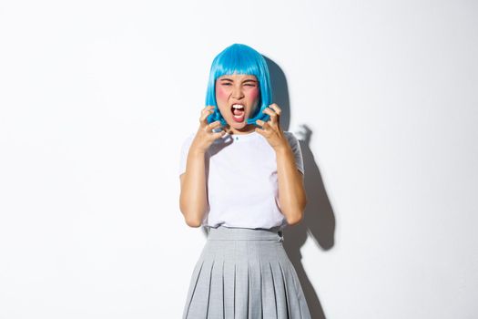 Portrait of angry asian girl in blue wig, yelling at someone with hatred, standing over white background.