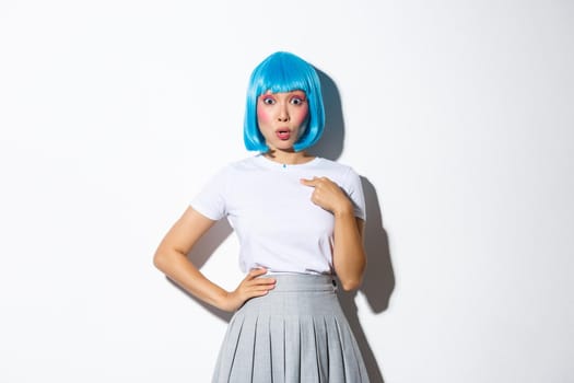 Image of surprised asian girl gasping amazed and pointing finger at herself, wearing blue wig for halloween party, standing over white background.