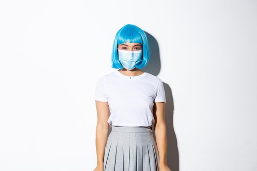 Concept of halloween during coronavirus. Image of cute asian girl in blue wig looking at camera, wearing medical mask, standing over white background.