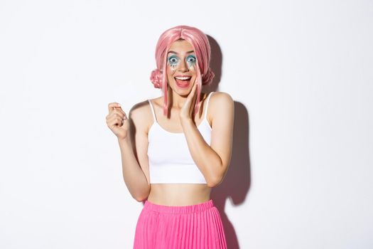 Cheerful pretty girl with bright makeup, dressed for holiday celebration or party, showing credit card and smiling excited, standing over white background.