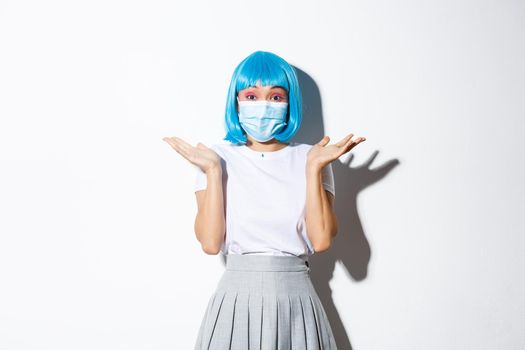 Concept of halloween celebration and covid-19 pandemic. Image of cute asian girl having fun, wearing blue wig and medical mask, shrugging clueless, standing over white background.