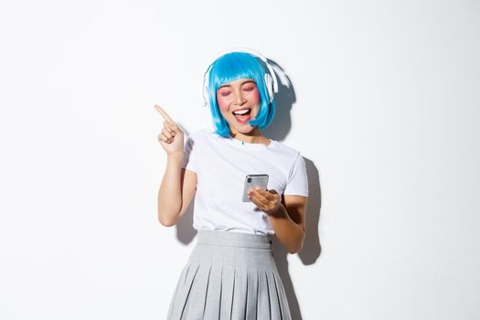 Image of pretty asian girl in blue party wig, listening music in headphones, singing along, holding smartphone.