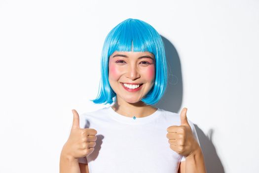 Close-up of happy asian girl in blue wig, smiling and showing thumbs-up in approval, like something good, standing over white background.