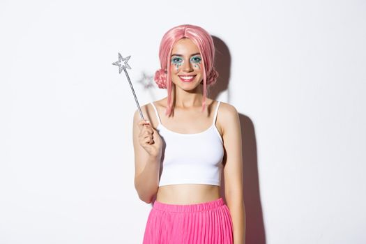Image of beautiful girl dressed-up as a fairy for halloween party, holding magic wand and smiling, standing over white background.