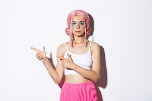 Image of beautiful caucasian girl in pink party wig and bright makeup pointing fingers left at your logo, showing banner about holidays or halloween, standing over white background.