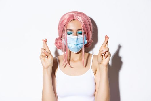 Coronavirus, social distancing and lifestyle concept. Close-up of hopeful girl in pink wig and medical mask cross fingers and making wish.