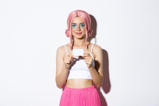 Portrait of cute smiling party girl in pink wig, showing credit card, standing over white background.