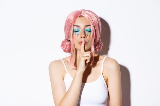 Close-up of lovely girl with pink wig and bright makeup, close eyes and shushing, have surprise or secret, standing over white background.