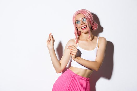 Image of attractive party girl with pink wig and bright makeup, having fun and celebrating holiday, dancing happy over white background.