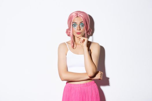 Excited cute party girl with pink wig and halloween makeup, looking with interest at camera, listening to something, standing over white background.