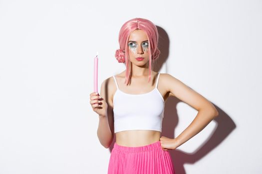 Image of thoughtful attractive girl in halloween costume and wig, holding pink candle and looking left while thinking, standing over white background.