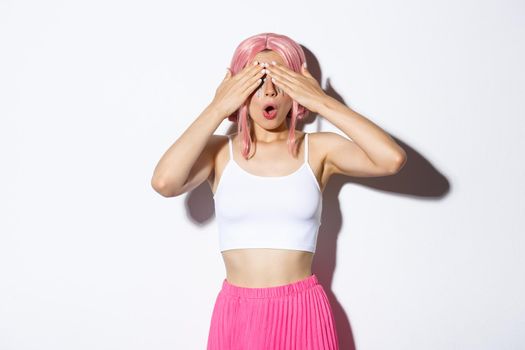 Portrait of intrigued cute girl with pink hair, close eyes with hands and open mouth wondered, waiting for surprise gift on holiday celebration, standing over white background.