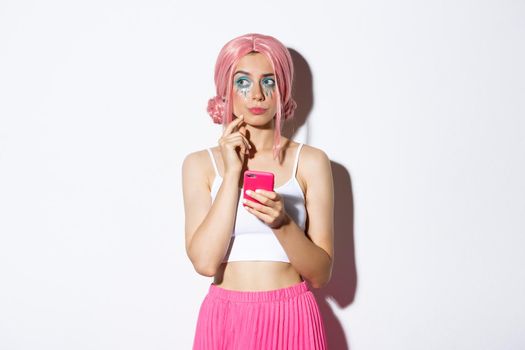 Portrait of thoughtful young woman in pink wig, with bright makeup, looking aside while thinking, holding mobile phone, standing over white background.